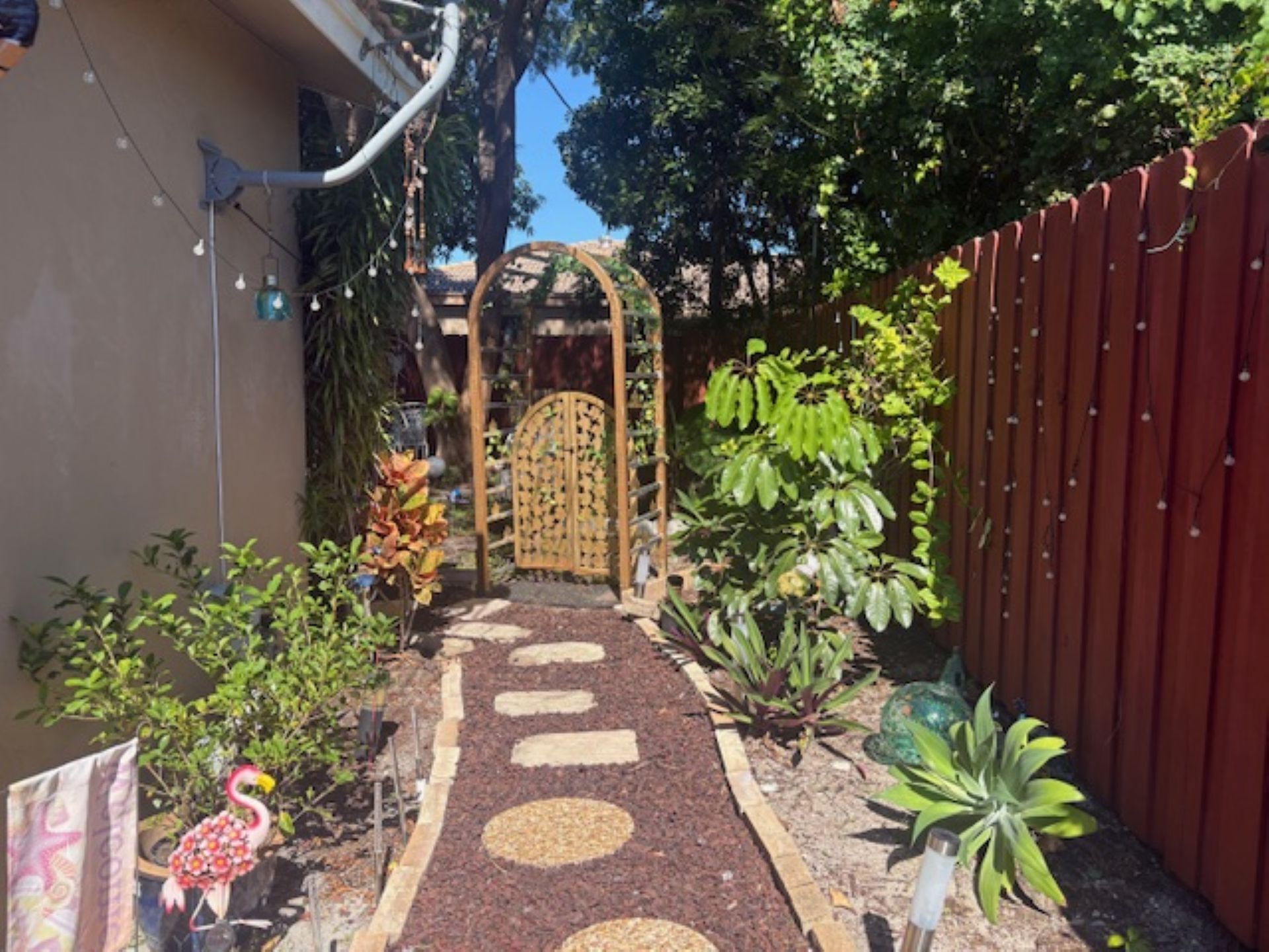 Un sendero en un jardín que lleva a una puerta de madera, rodeado de plantas variadas y una valla roja.