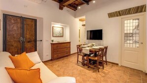 Ein Zimmer mit Holzboden, einem Esstisch für vier Personen, einer Couch mit orangefarbenen Kissen und einem an der Wand befestigten Fernseher.