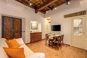 Ein Zimmer mit Holzboden, einem Esstisch für vier Personen, einer Couch mit orangefarbenen Kissen und einem an der Wand befestigten Fernseher.
