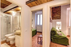 Un baño con ducha, inodoro y lavamanos, un sofá verde en la sala de estar, y techo de madera.