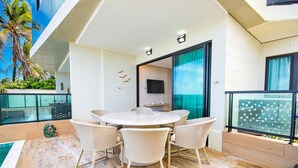 A modern outdoor dining area with a round table and wicker chairs, a TV mounted on the wall, and a view of palm trees. - Vila Porto by Carpediem In Front of Porto de Galinhas Beach (Ipojuca)