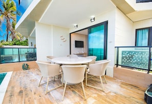 A modern outdoor dining area with a round table and wicker chairs, a TV mounted on the wall, and a view of palm trees. - Vila Porto by Carpediem In Front of Porto de Galinhas Beach (Ipojuca)