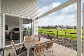 Eine überdachte Terrasse mit Tisch und Stühlen, eine Glasschiebetür zu einem Innenbereich und Blick auf ein grünes Feld.