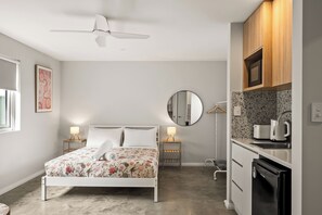 Ein modernes Schlafzimmer mit einem Bett, Nachttischlampen, einem Deckenventilator, einem runden Spiegel und einer Küchenzeile mit einer Kaffeemaschine.