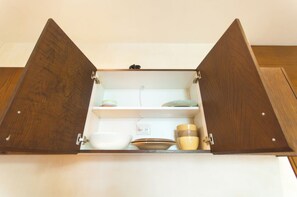Ein Holzkabinent mit zwei Türen, eine davon geöffnet, zeigt ein weißes Regal mit einer Schüssel, einem Teller und einer Tasse.
