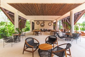 An outdoor seating area with wicker chairs and tables, surrounded by greenery and a covered roof structure. - Cozy 1-bedroom condo in serene Tulum with AC, pool, gym access and bikes (Tulum)