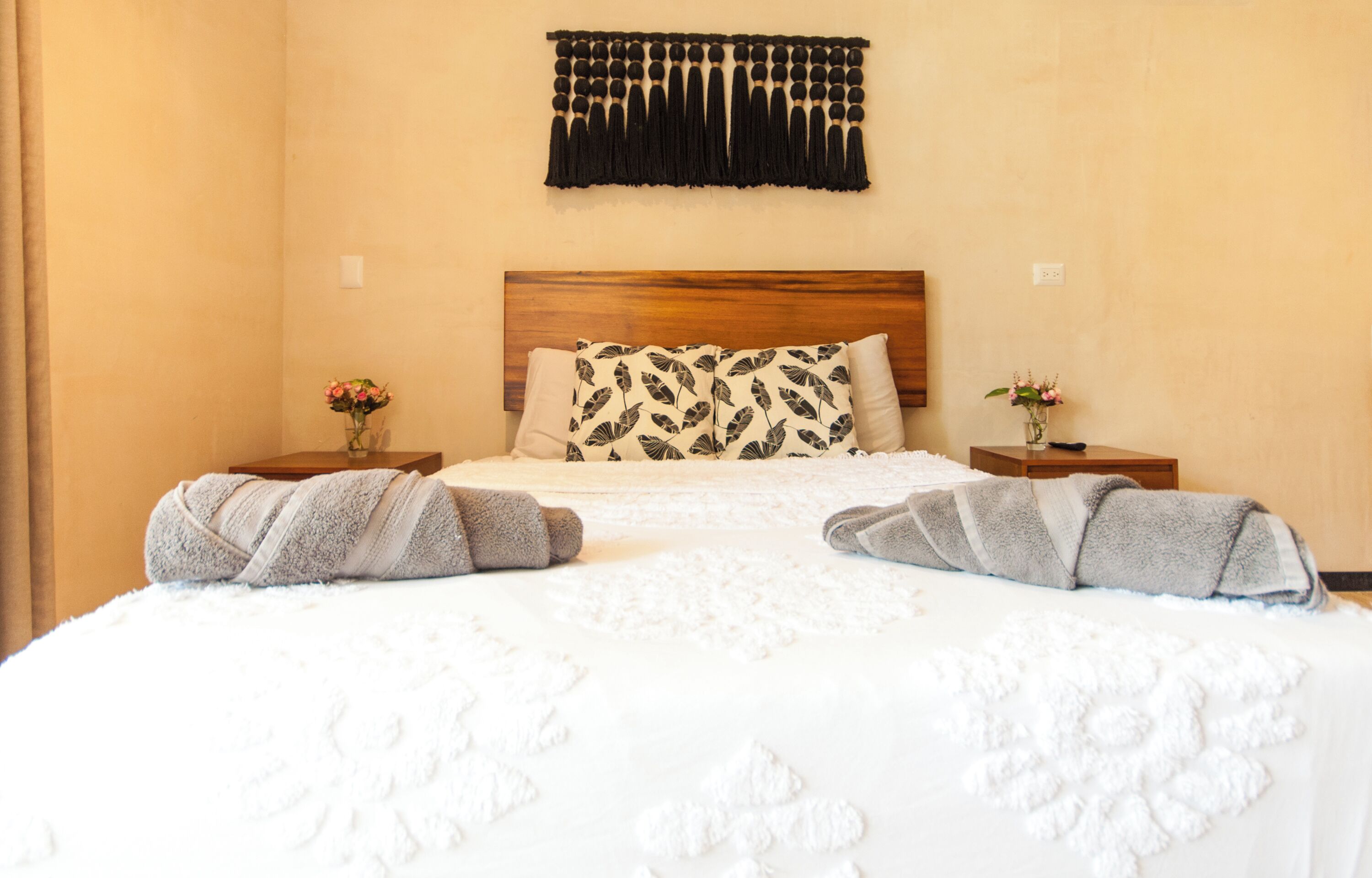 A hotel room with a bed, two nightstands, a wooden headboard, a wall-mounted tassel decoration, and two vases with flowers.