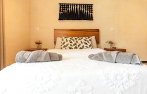 A hotel room with a bed, two nightstands, a wooden headboard, a wall-mounted tassel decoration, and two vases with flowers. - Cozy 1-bedroom condo in serene Tulum with AC, pool, gym access and bikes (Tulum)