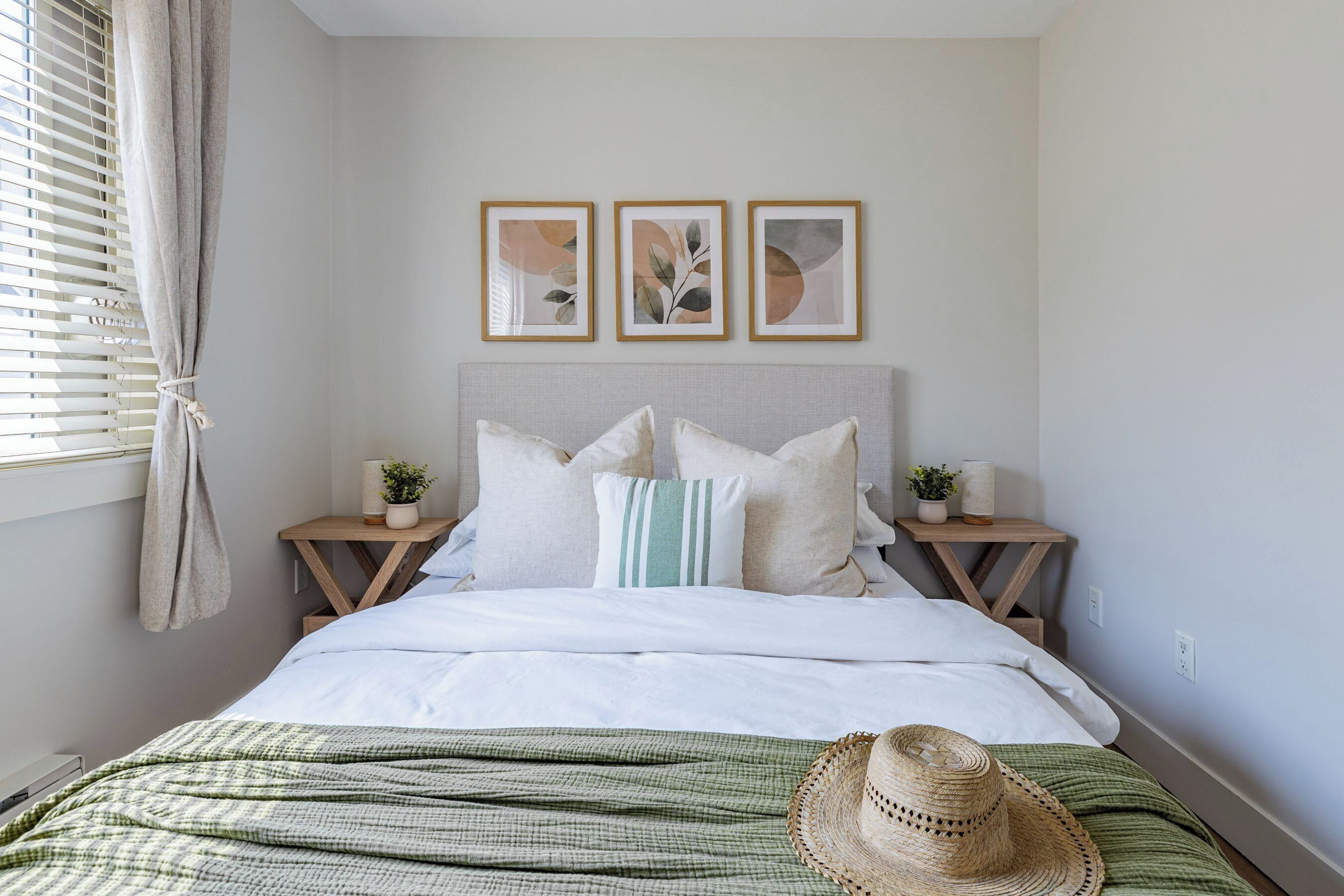 Een slaapkamer met een bed, kussens, een nachtkastje met een lamp, een plant en een aan de muur hangend fotolijstje.