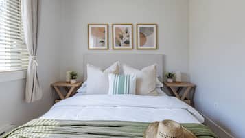 Een slaapkamer met een bed, kussens, een nachtkastje met een lamp, een plant en een aan de muur hangend fotolijstje.