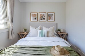 A bedroom with a bed, pillows, a bedside table with a lamp, a plant, and a wall-mounted picture frame. - Suburban Escape - Coach House (Surrey)