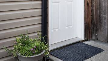 A white door with a wreath, a potted plant with purple flowers, and a black mat at the entrance.