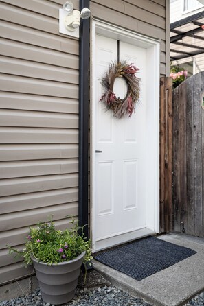 A white door with a wreath, a potted plant with purple flowers, and a black mat at the entrance. - Suburban Escape - Coach House (Surrey)