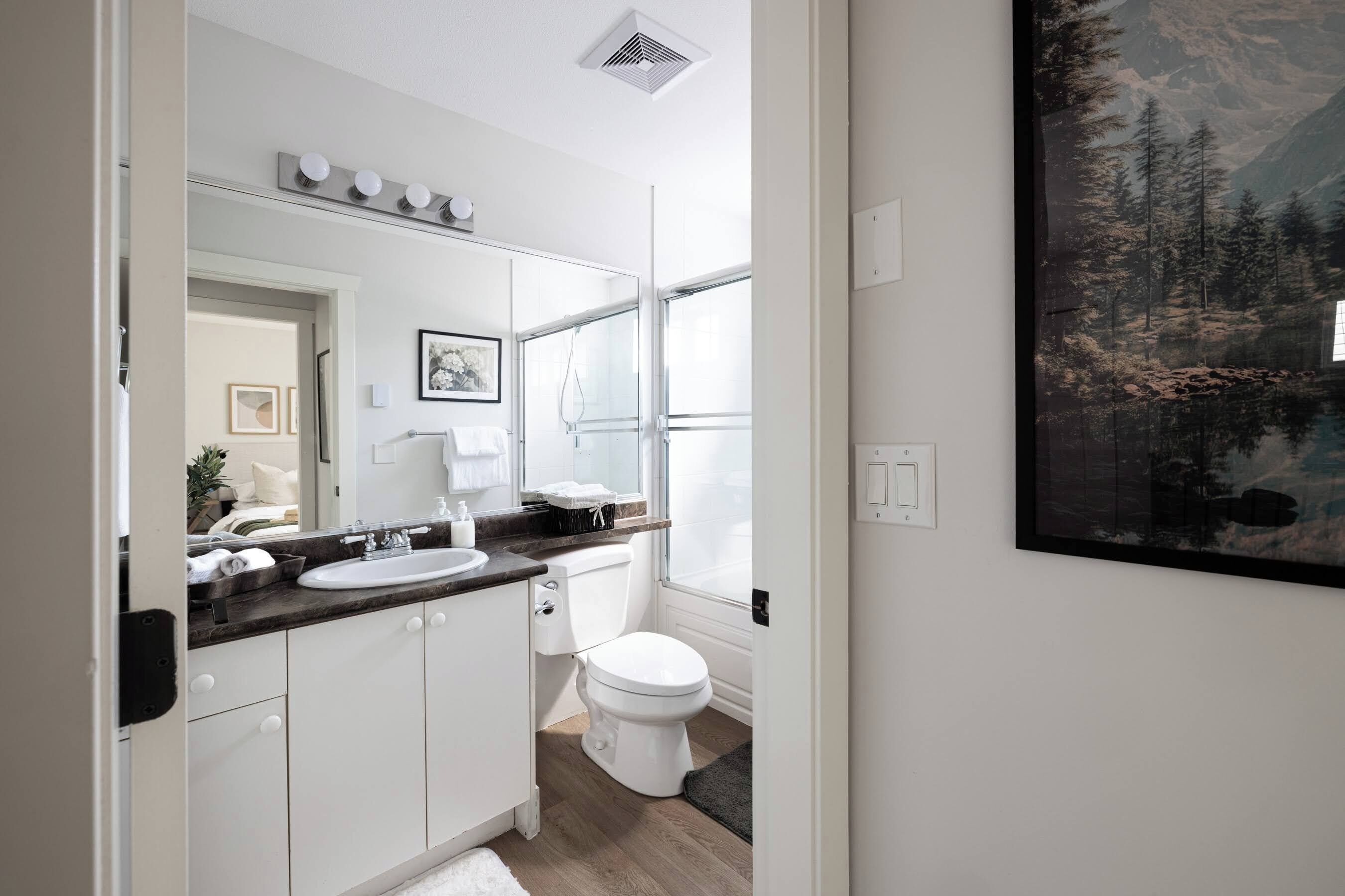 A modern bathroom with a white sink, mirror, and toilet. There is a window with a view of trees and a painting on the wall.