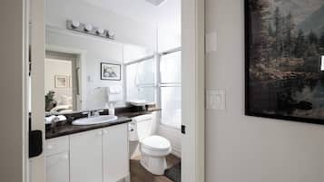 A modern bathroom with a white sink, mirror, and toilet. There is a window with a view of trees and a painting on the wall.