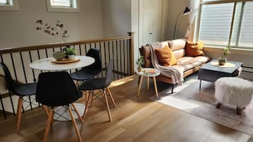 Een kamer met een eethoek, bestaande uit een ronde tafel en stoelen, een bank met kussens, een klein bijzettafeltje met een lamp, en een raam met jaloezieën.