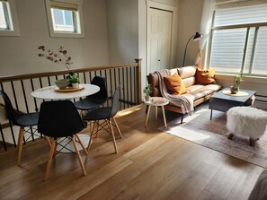 A room with a dining area featuring a round table and chairs, a sofa with cushions, a small side table with a lamp, and a window with blinds. - Suburban Escape - Coach House (Surrey)