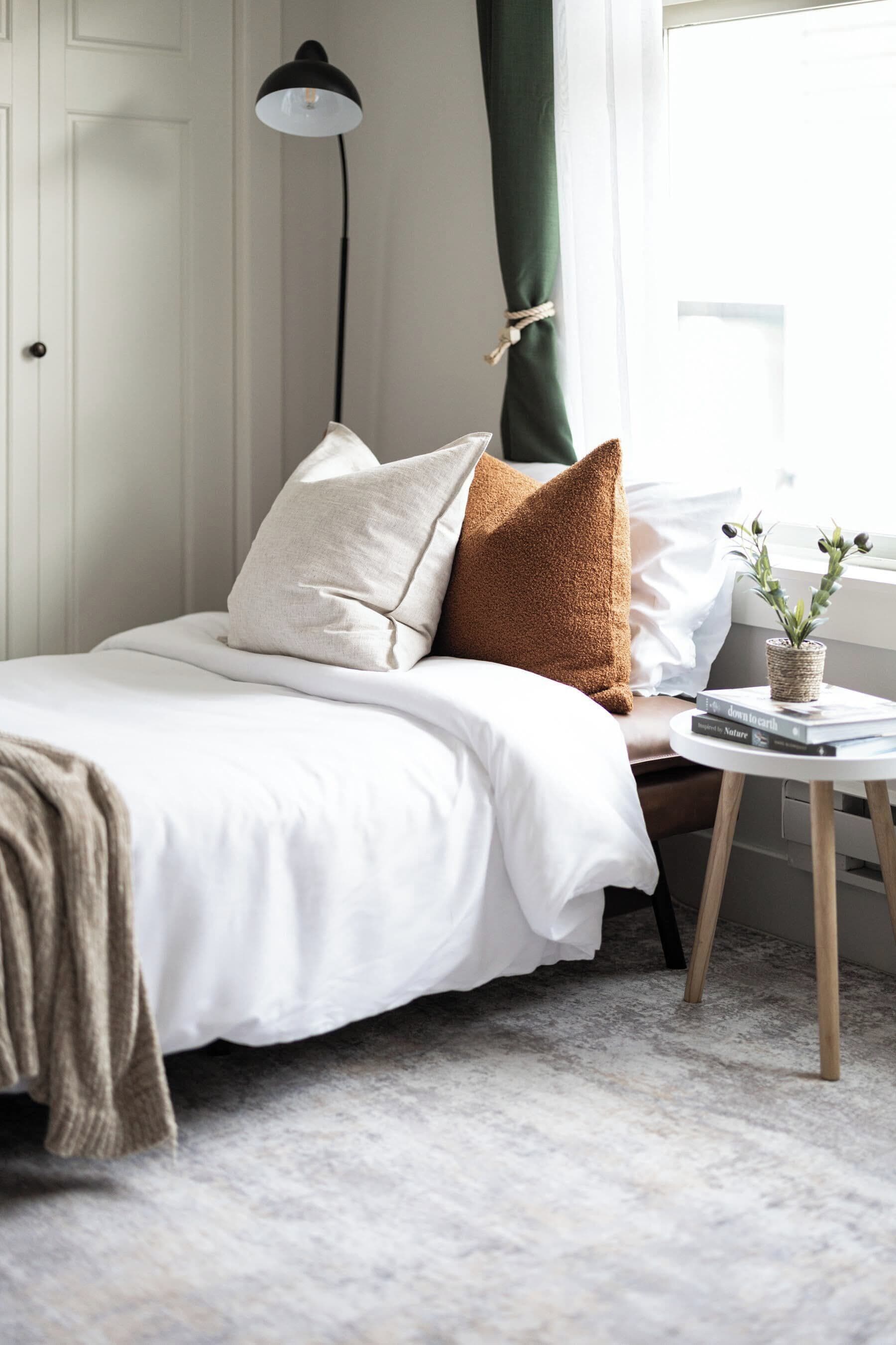 Una cama bien tendida con una colcha blanca y dos almohadas, una pequeña mesa redonda con una planta y libros, y una lámpara de pie.