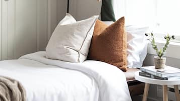 Una cama bien tendida con una colcha blanca y dos almohadas, una pequeña mesa redonda con una planta y libros, y una lámpara de pie.