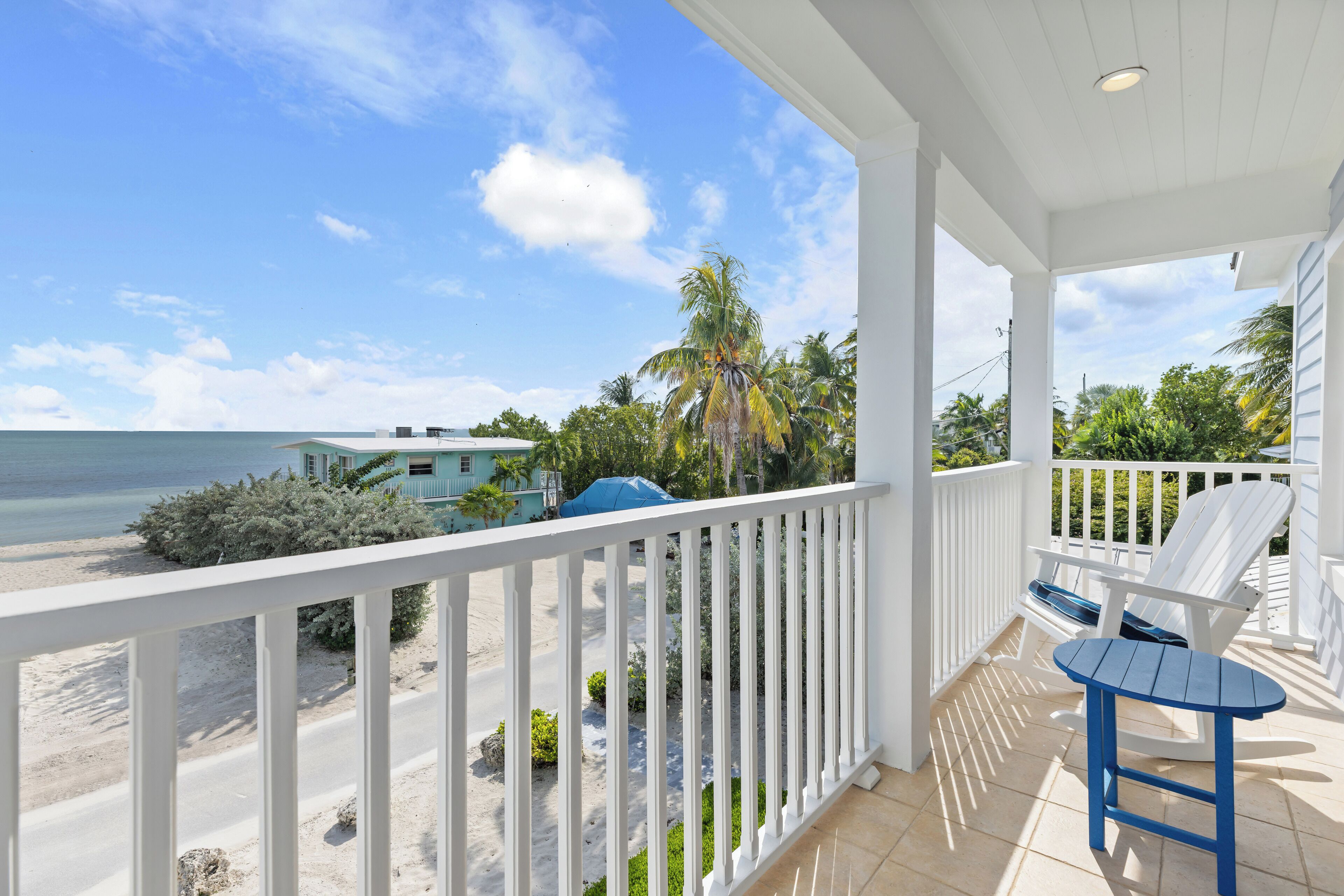 A balcony with a view of the beach and ocean, white railings, and a blue table and chair.