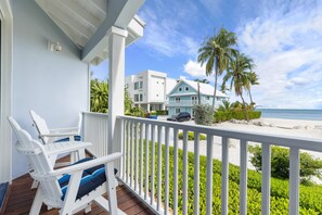 A balcony with white railings, a white chair with blue cushions, and a view of a beach with palm trees and a house. - Islamorada Beach Retreat. Private Beach, Pool, and Boat Dock! (Islamorada)