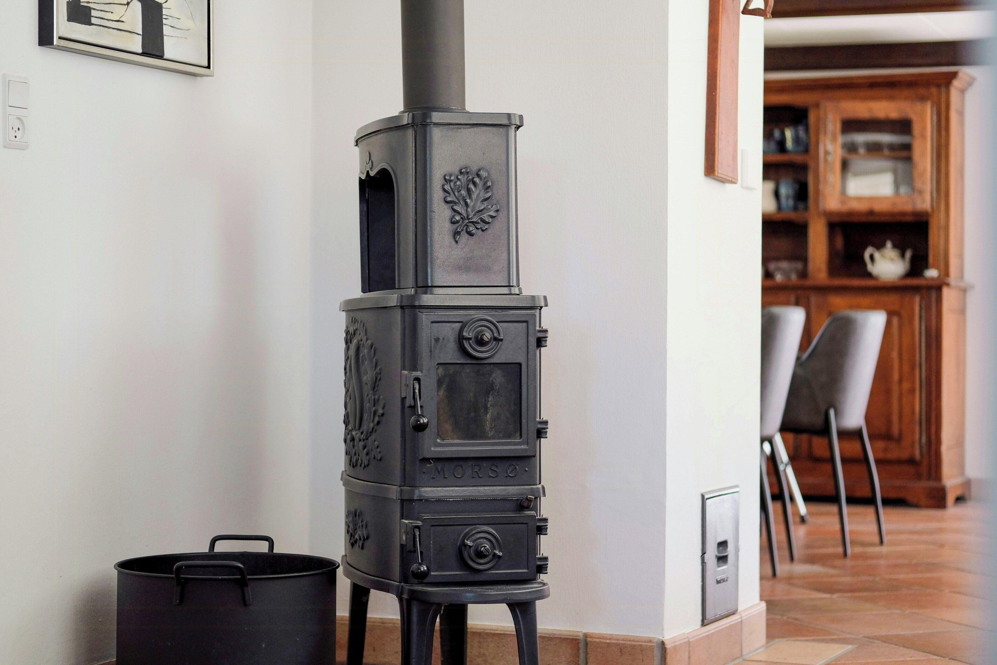 A black wood-burning stove with a chimney, a black pot, and a chair in a room with a tiled floor and a wooden cabinet in the background.