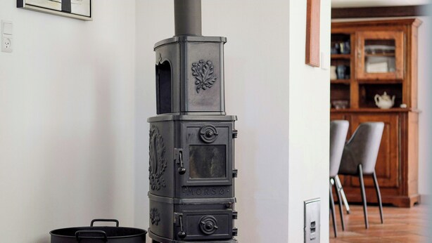 Una estufa de leña negra con chimenea, una olla negra y una silla en una habitación con piso de baldosas y un armario de madera al fondo.