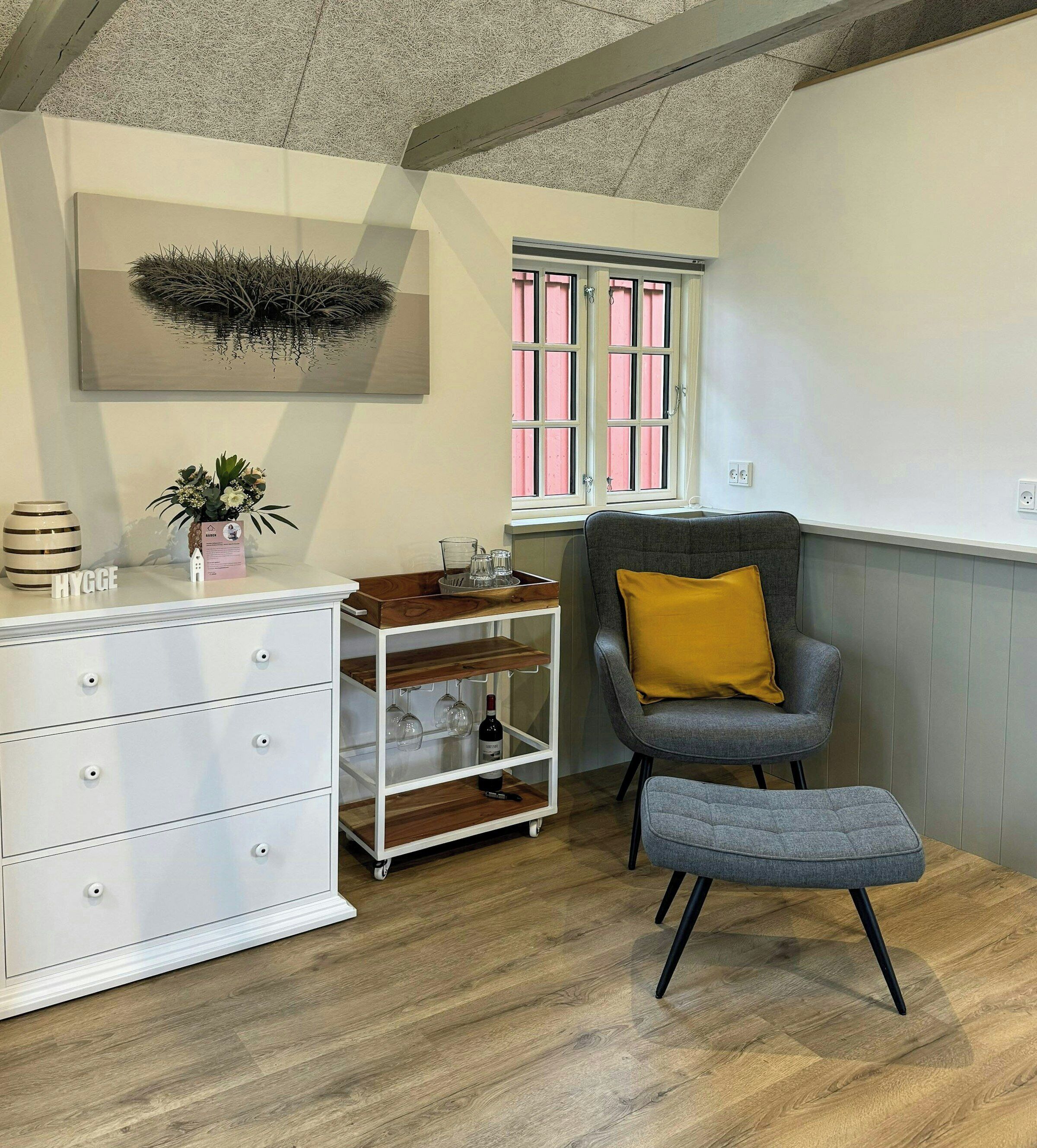 A modern room with a white dresser, a grey armchair with a yellow pillow, a small wooden table, and a wall-mounted artwork.