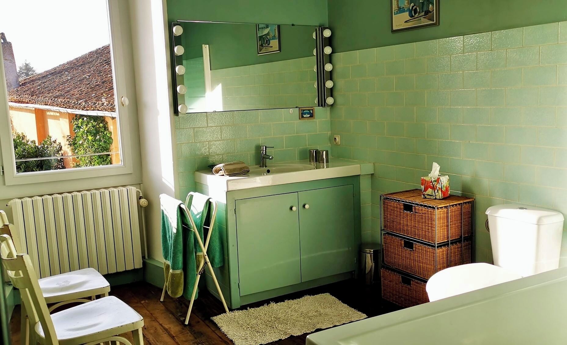 A bathroom with a green tiled wall, a vanity with a mirror and lights, a radiator, a wicker basket, and a window with a view of a building outside.