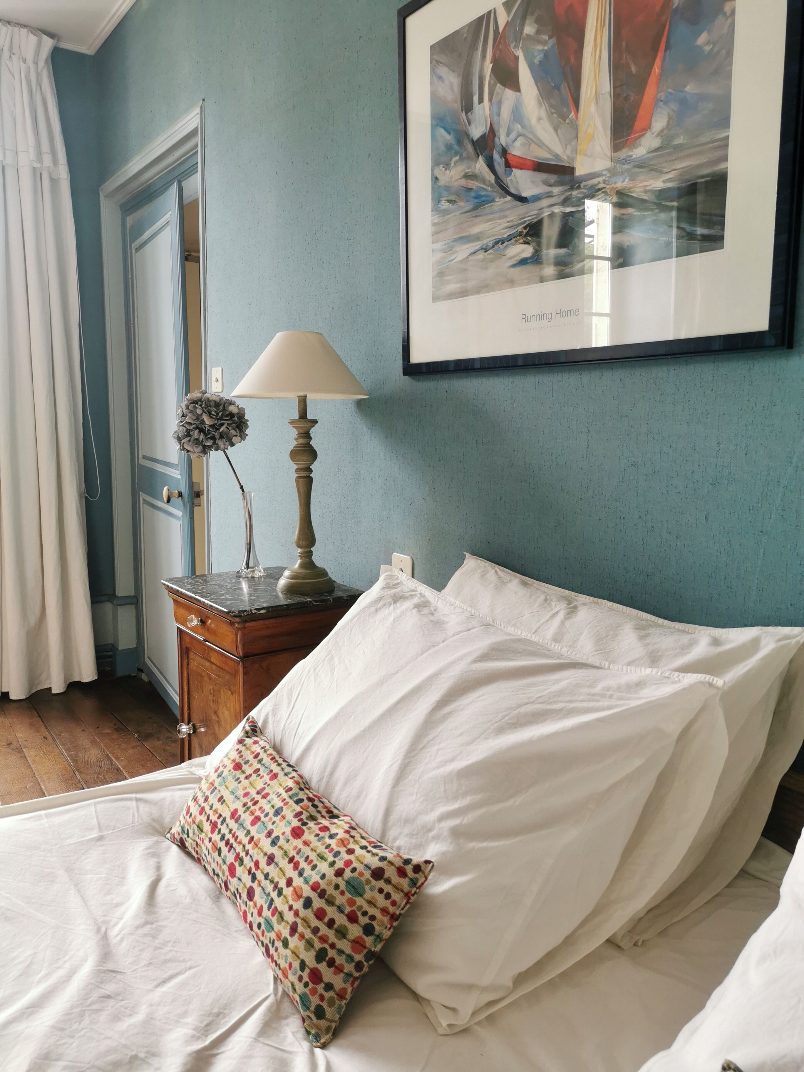 A bedroom with a bed, pillows, a nightstand with a lamp, a door, and a framed picture on the wall.