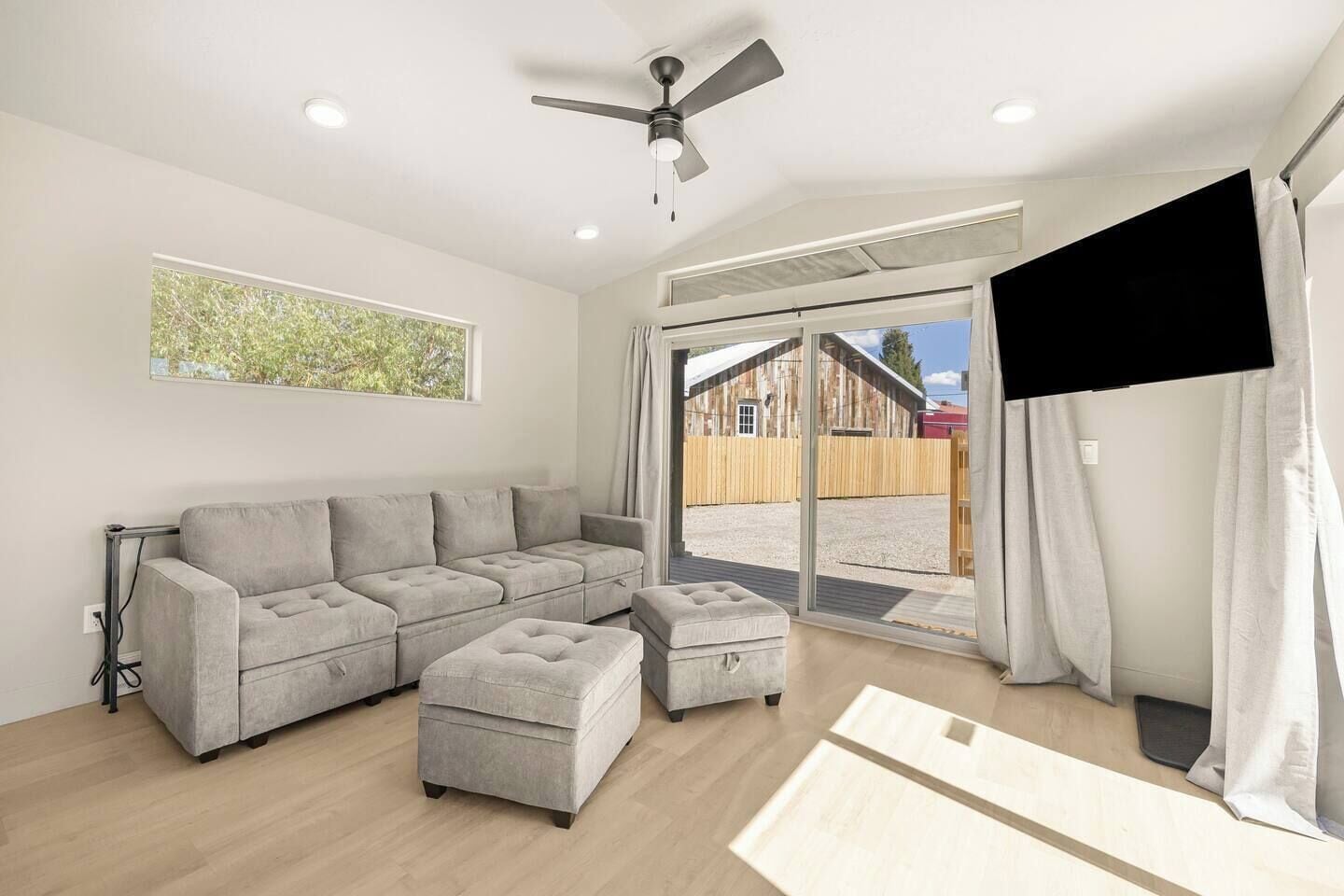 Ein modernes Wohnzimmer mit Ecksofa, Deckenventilator und Blick auf einen hölzernen Bauernhof durch eine Schiebetür.