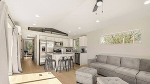 Una cocina moderna con una isla central, taburetes de bar y un sofá gris en una sala de estar con vista a árboles.