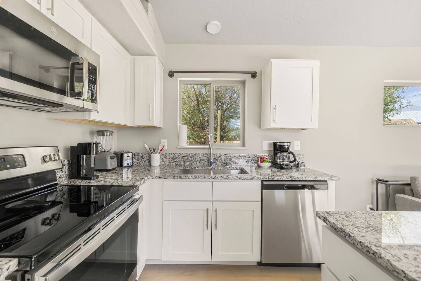 A modern kitchen with white cabinetry, a granite countertop, a built-in oven, a dishwasher, and a window with a view of trees.