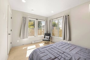 Un dormitorio con cama, una silla y dos ventanas con cortinas.
