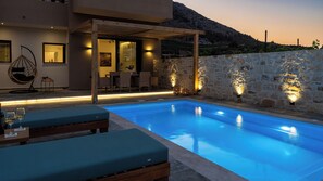A modern outdoor pool area with a stone wall and wooden pergola, surrounded by lounge chairs and a dining area. - Irisma Luxury Villa I | Private Pool near Heraklion, Crete (Archanes)