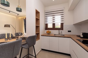 A modern kitchen with a dining area, featuring a wooden table, grey chairs, a fruit bowl, and a window with blinds. - Irisma Luxury Villa I | Private Pool near Heraklion, Crete (Archanes)