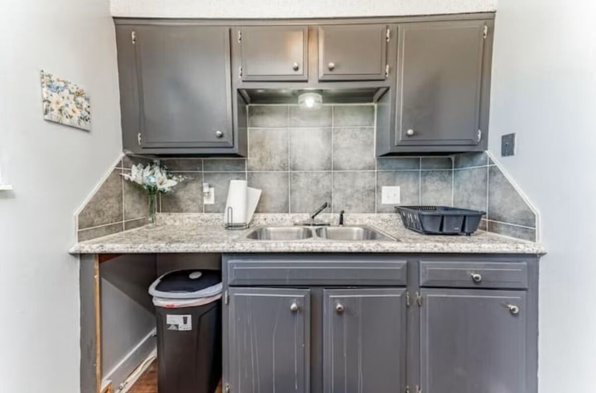 A compact kitchen with grey cabinets, a granite countertop, a sink, and a trash bin.