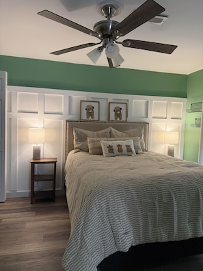 A bedroom with a bed, two bedside lamps, a ceiling fan, and a wall-mounted shelf with framed pictures.