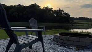 Zona relax con due poltrone Adirondack su fondo ghiaioso, un braciere in pietra e vista su un lago con alberi sullo sfondo.