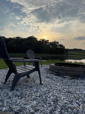Ein Sitzbereich mit zwei Adirondack-Stühlen auf einer Schotterfläche, einem Steinkamin und Blick auf einen See mit Bäumen im Hintergrund.