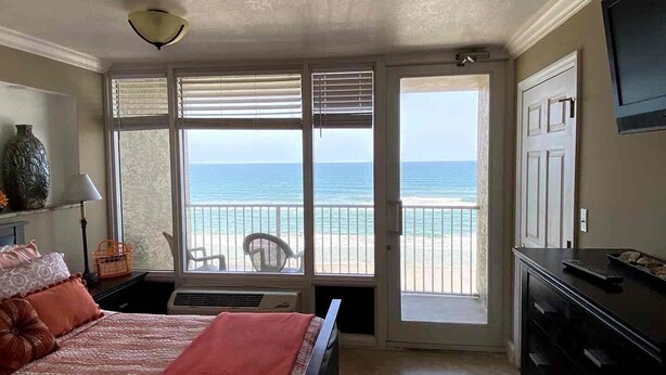 Ein Schlafzimmer mit einem Bett, einer Kommode, einer Lampe, einer Vase und Blick auf Strand und Meer durch große Fenster.