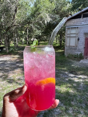 Una mano sostiene un vaso alto con pajita, lleno de una bebida rosa y hielo, al aire libre, con árboles y un edificio de madera al fondo.
