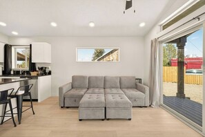 Un espacio habitable moderno con un sofá seccional gris, una zona de cocina con armarios blancos y un amplio ventanal que ofrece vistas al exterior.