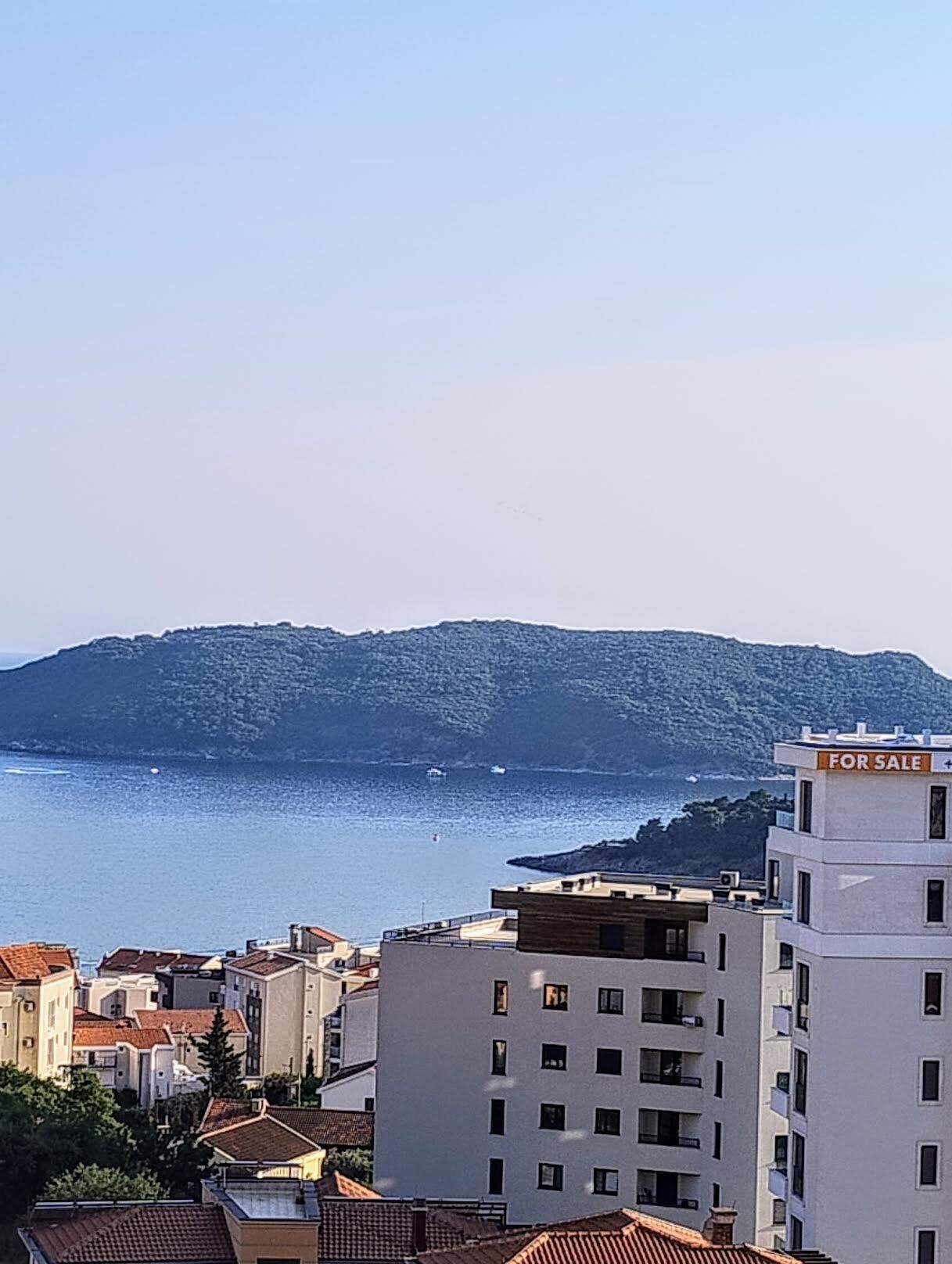 A coastal cityscape with buildings, a 'For Sale' sign, and a body of water.