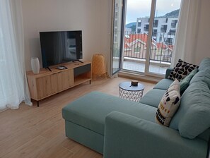 A modern living room with a sofa, a TV on a wooden stand, a small round table, and a view of buildings through a glass door. - Cozy 1-bedroom apartment in lovely Budva with sea view. (Budva)