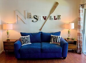 Ein blaues Sofa mit zwei Kissen, eine Wand mit dem Schriftzug 'NASHVILLE' und einem Schläger, sowie zwei Lampen auf Beistelltischen.
