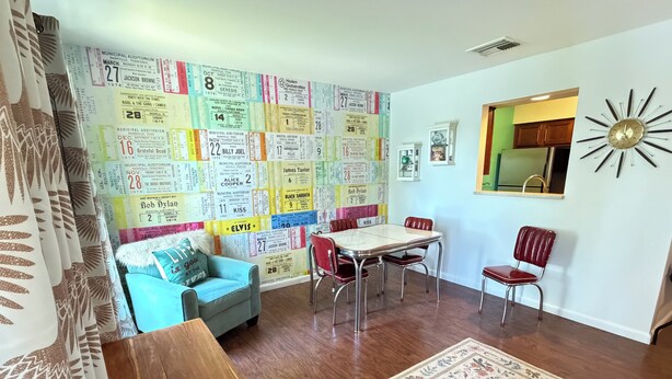 Ein Wohnzimmer mit einer farbenfrohen Wand aus Tickets, einem blauen Sofa, einem Esstisch mit Stühlen, einem roten Sessel und einer hölzernen Kommode.