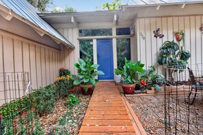 Une terrasse en bois menant à une porte bleue, entourée de plantes en pot et d’une clôture en métal.
