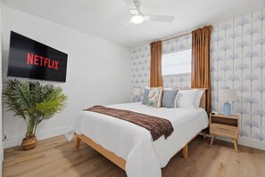 A bedroom with a bed, a TV mounted on the wall, a plant, and a window with curtains.