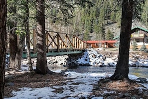 Eine hölzerne Brücke über einem Fluss, umgeben von Bäumen und einer Hütte mit rotem Dach.
